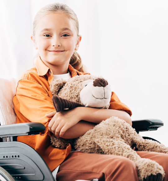 Ein pflegebedürftiges Kind im Rollstuhl mit einem Teddybär im Arm